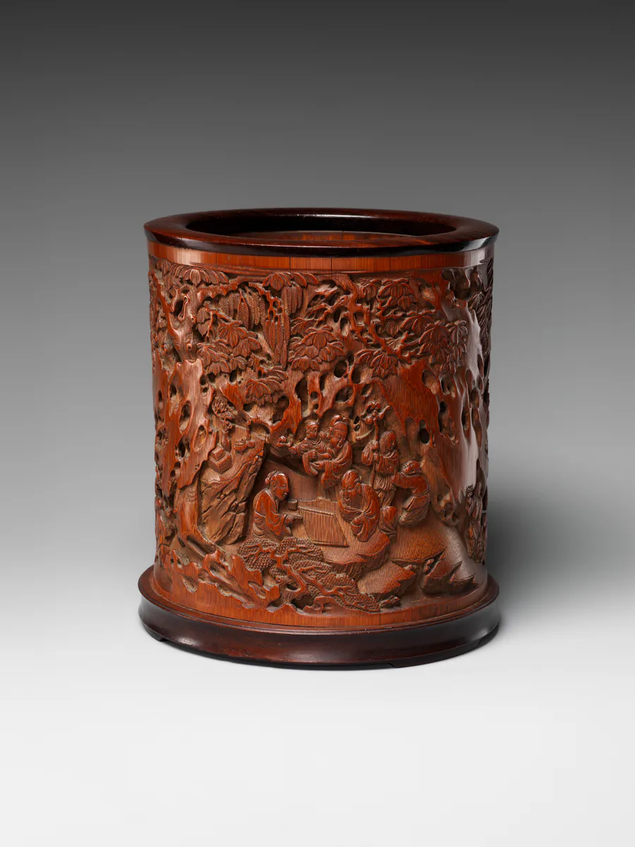 A cylindrical brush holder of red-brown bamboo, carved all round with figures among rocks and pines.